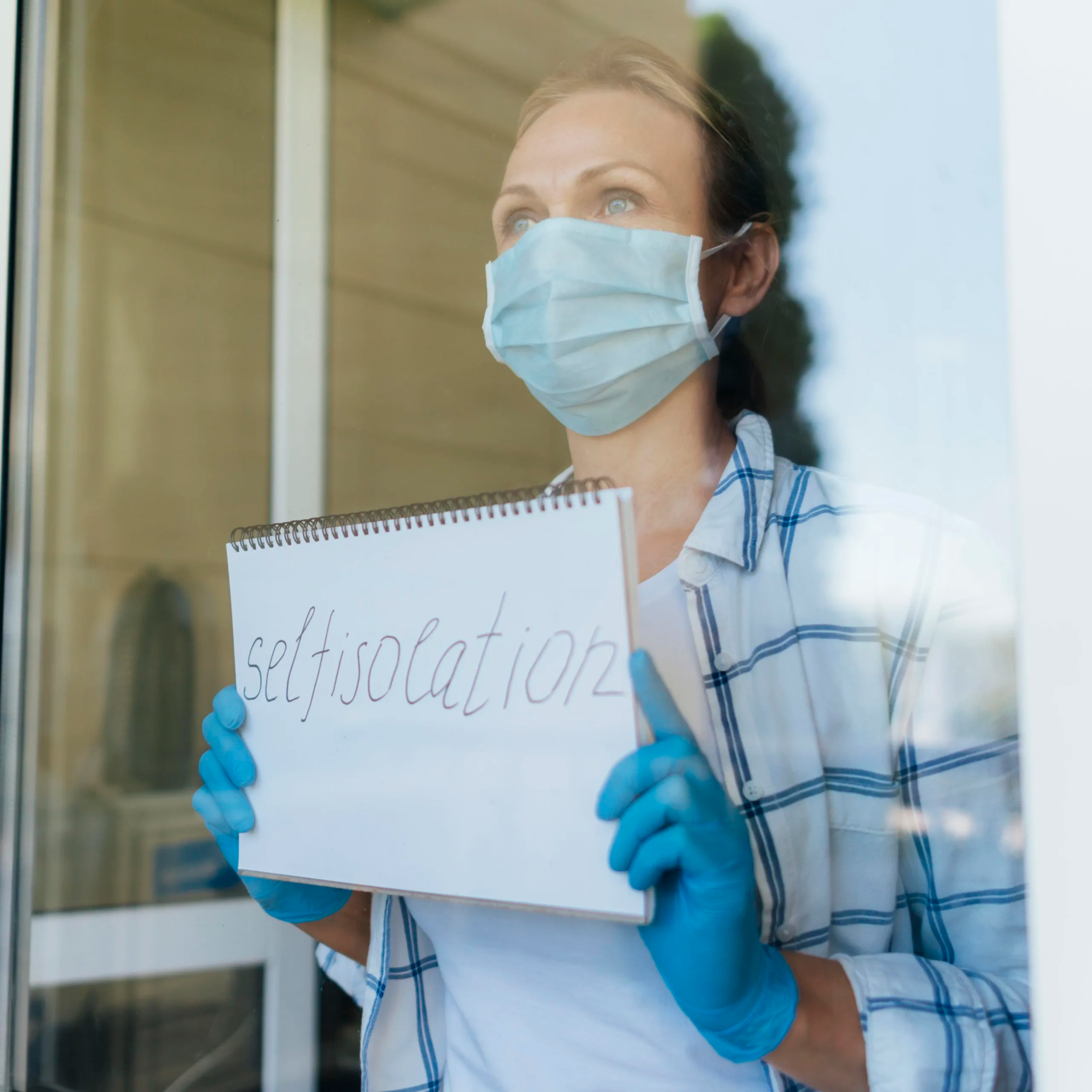 Vrouw met mondkapje en handschoenen kijkt door het raam en houdt een schrift met 'selfisolation' vast