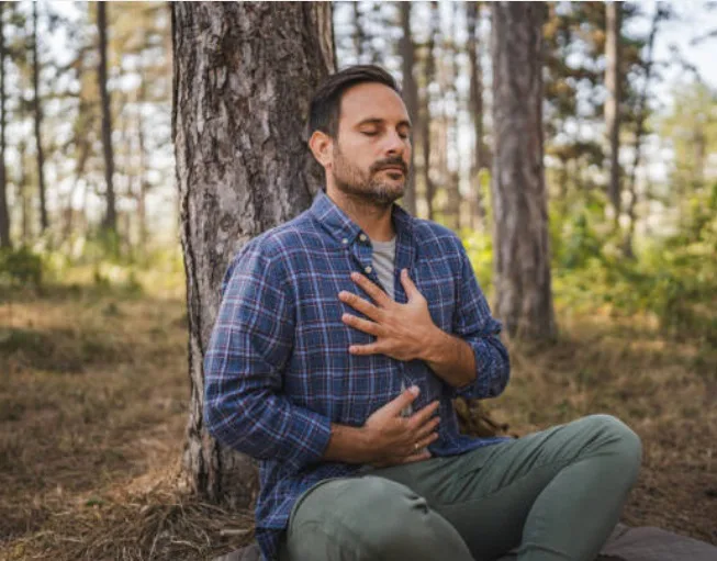 Man zit tegen een boom in het bos met zijn handen op borst en buik en ademt rustig