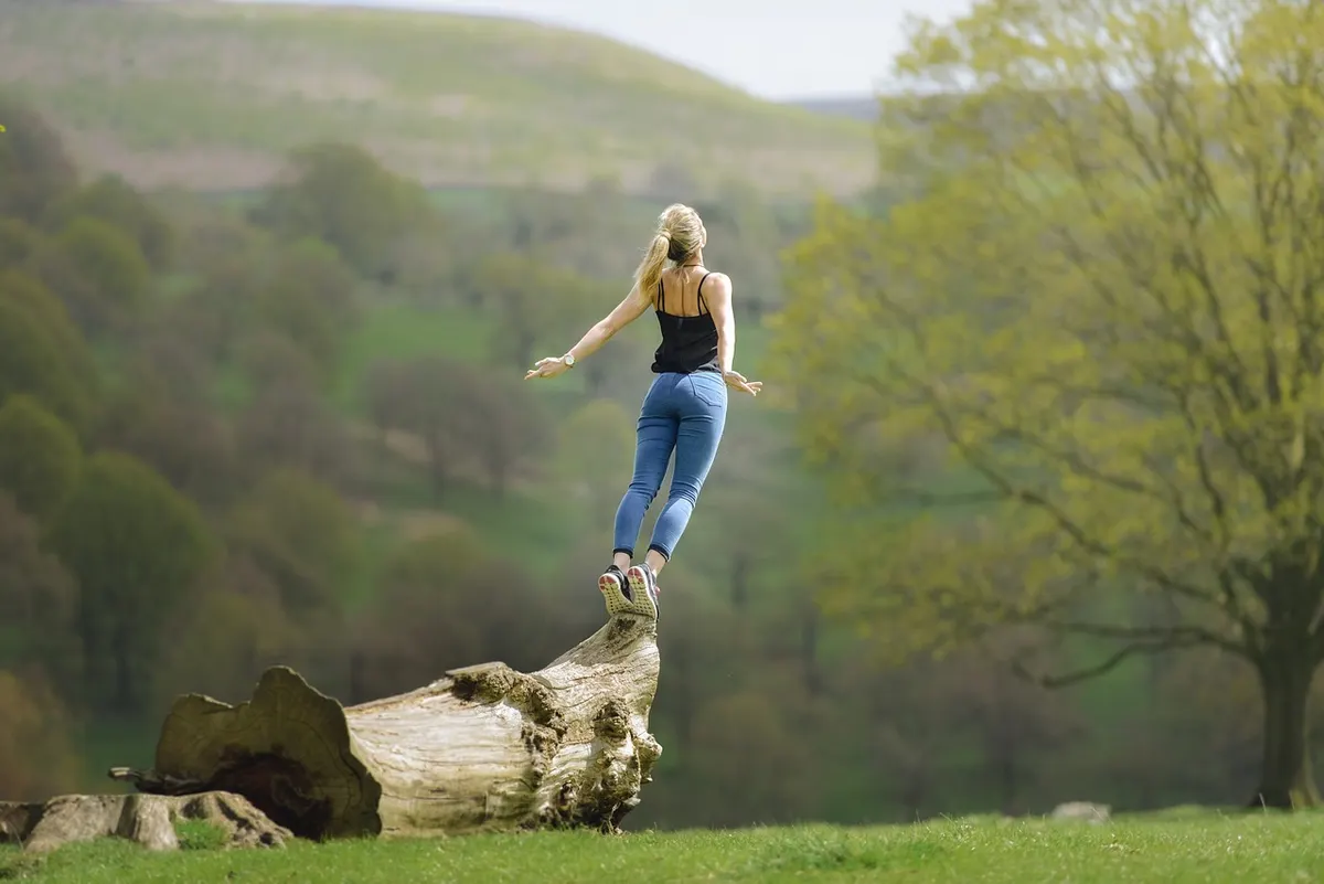 Vrouw springt met gespreide armen van een boomstam in een groen landschap